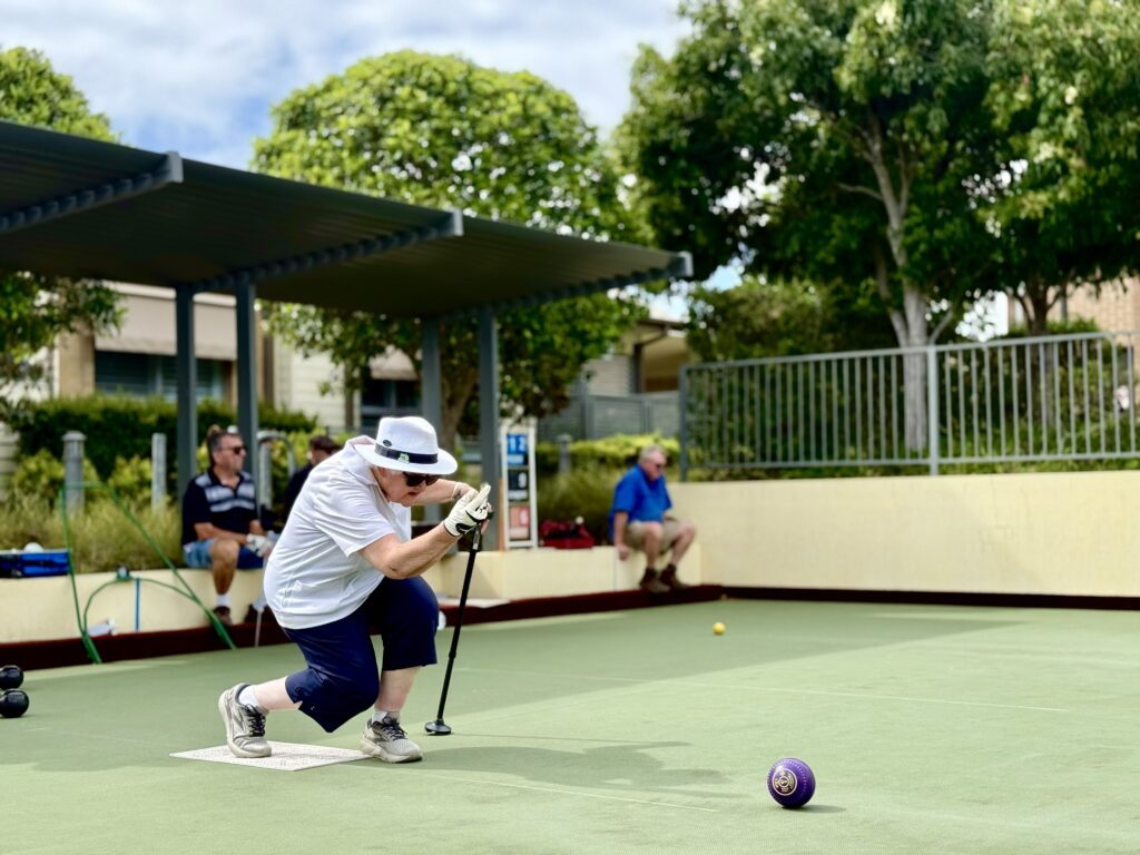 Kerry's Mum Joan enjoying a competitive game of lawn bowls at her retirement village. Joan is 88 years old, and has silver hair and is wearing a wide-brimmed hat, sunglasses, a white shirt and navy pants. Joan uses an adapted walking stick to help her balance as she releases her bowl. She has a competitive expression on her face.